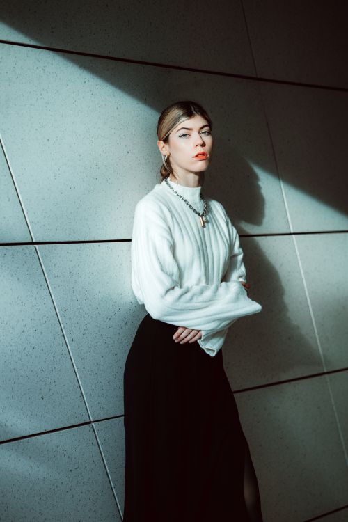 Free A fashionable woman stands elegantly against a modern wall, illuminated by sunlight. Stock Photo