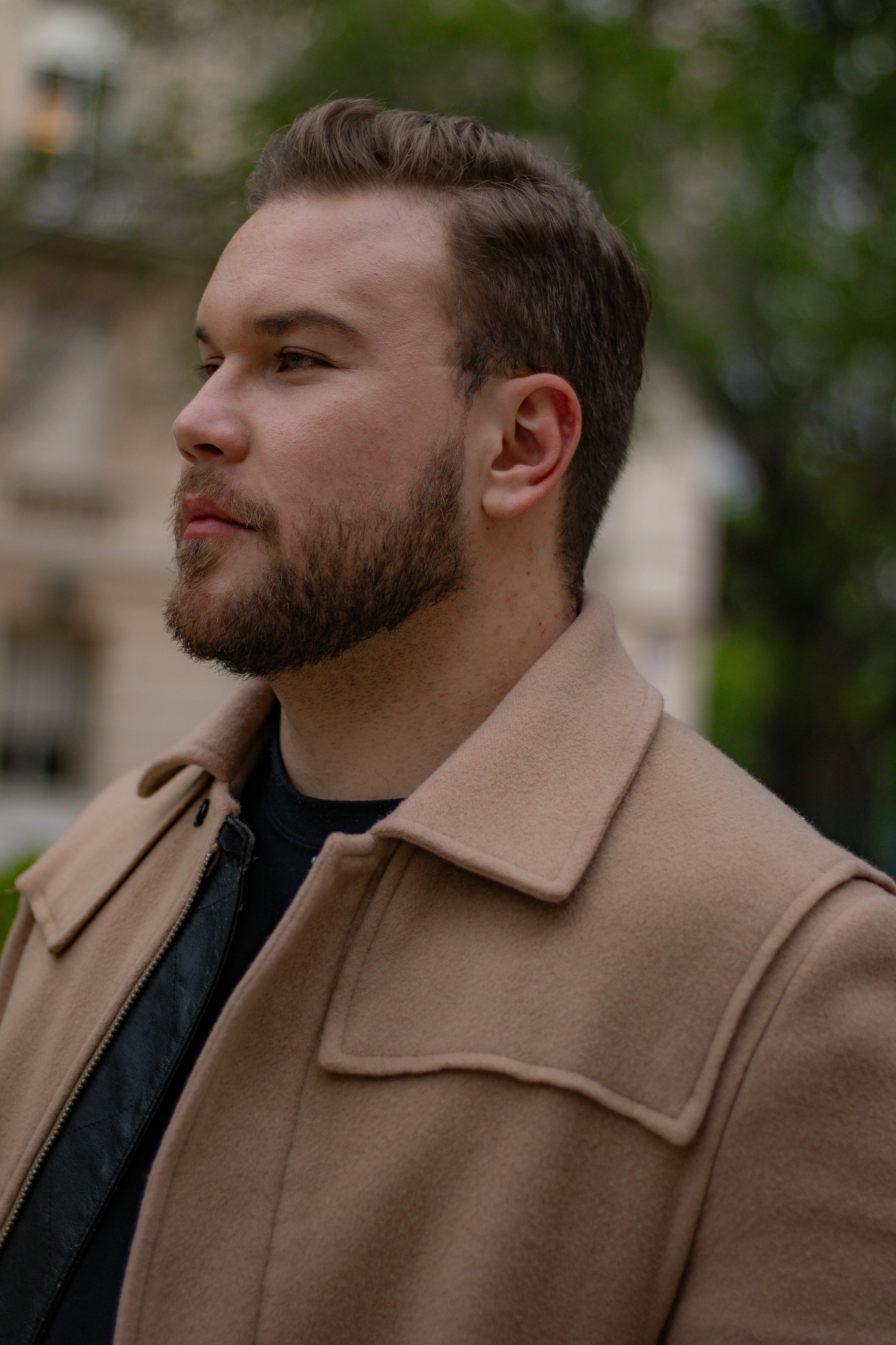 Profile portrait of a bearded man in a tan coat outdoors, showcasing modern fashion.