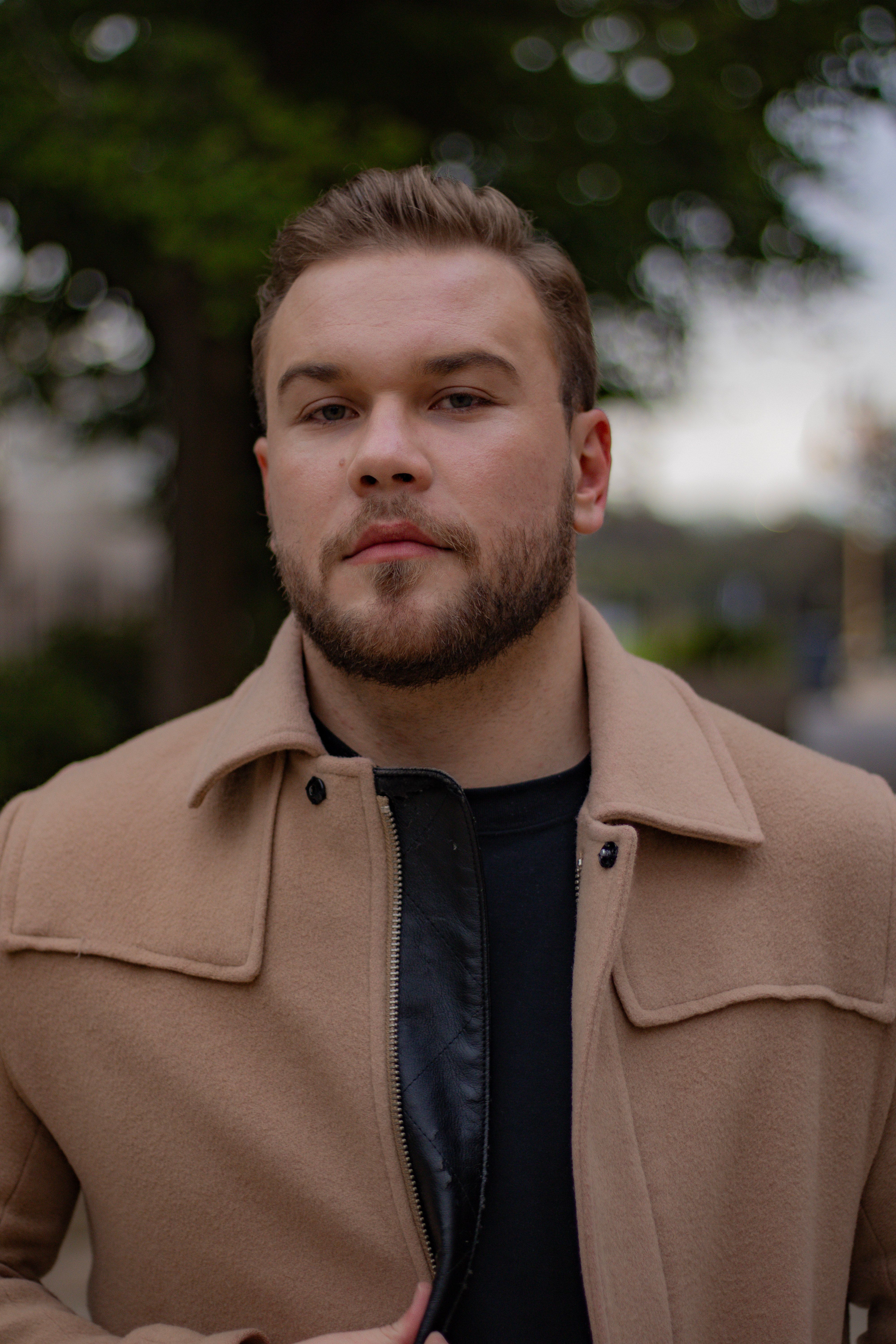 A fashionable adult man with a beard wearing a jacket, posed confidently outdoors.