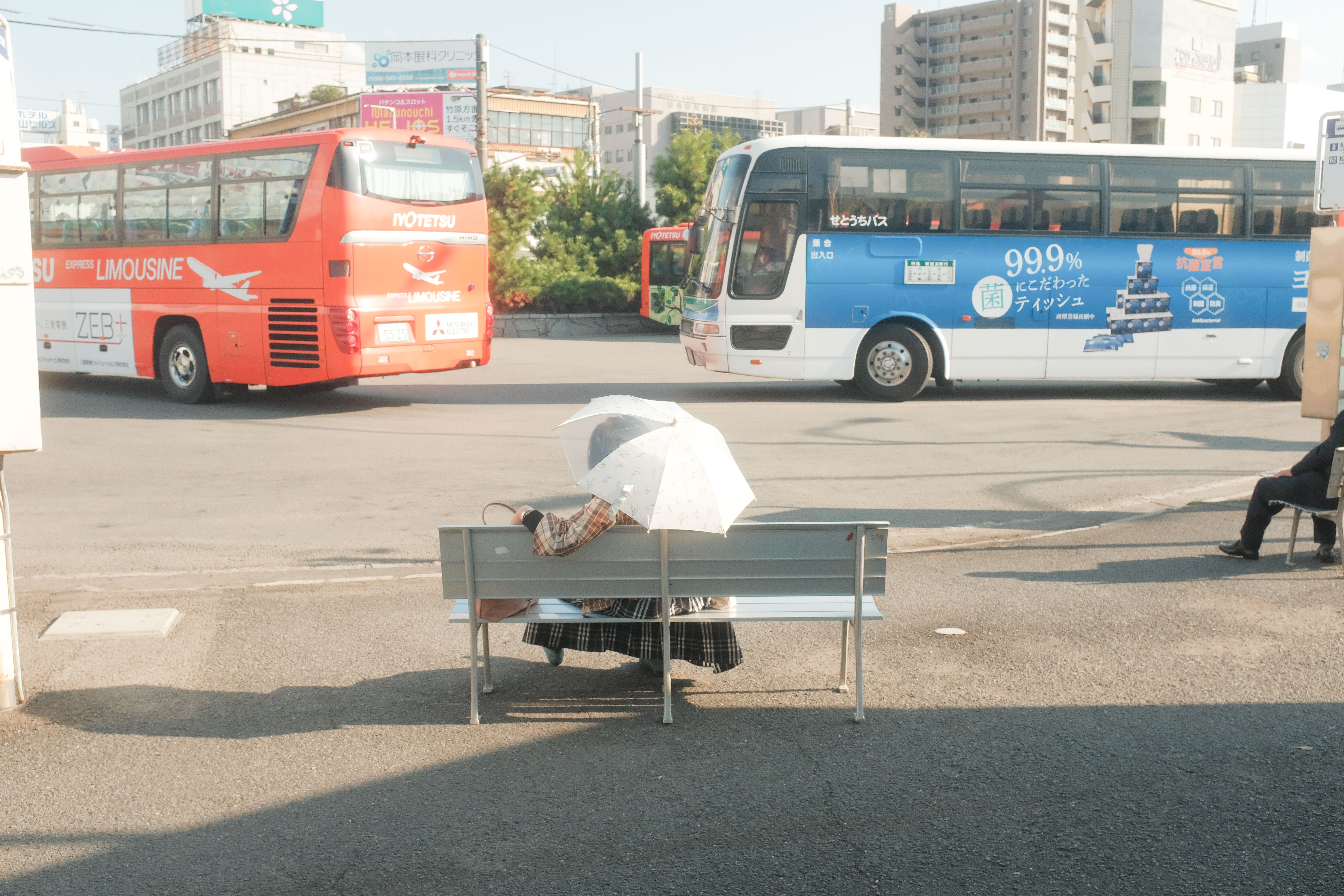 A woman sits on a bench with an umbrella at an urban bus stop, watching passing buses.