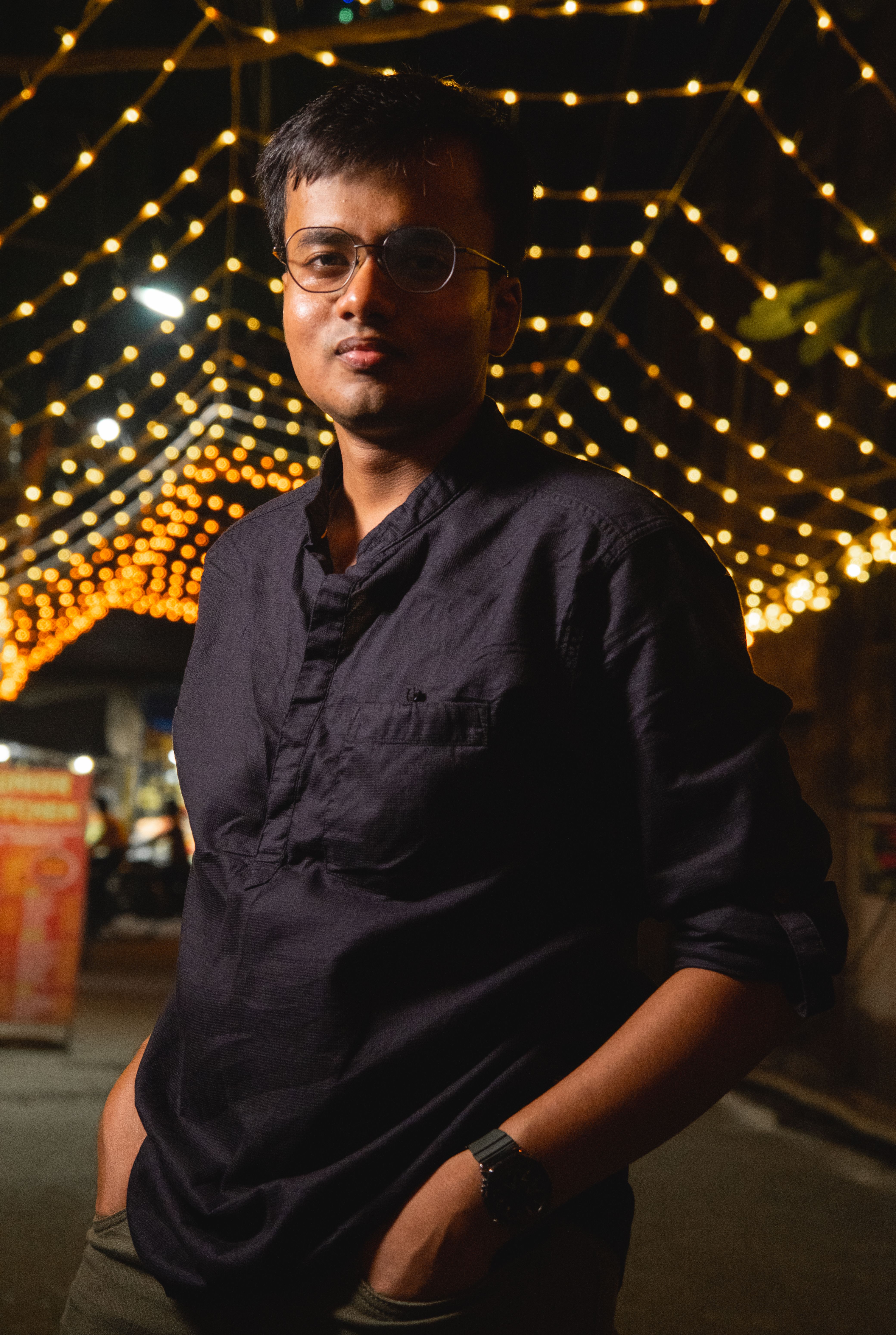 Portrait of a young man in a black shirt standing under glowing string lights in an urban setting at night.