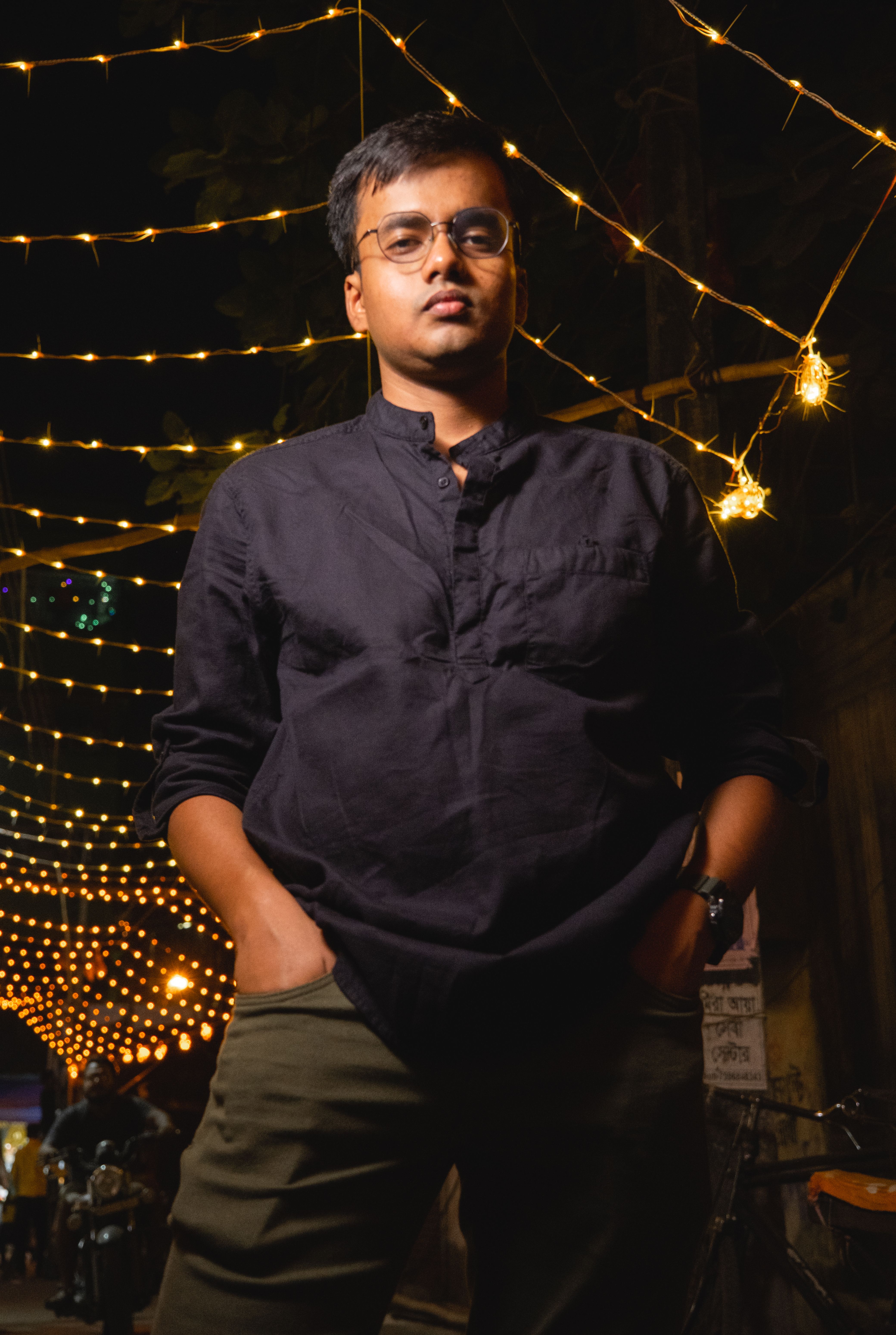 Young man in casual attire posing under festive string lights in a nighttime street setting.