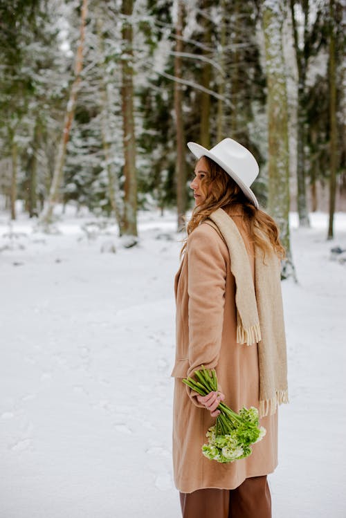 Woman in a Beige Coat and White Hat with a Bouquet of Flowers by the Winter Forest