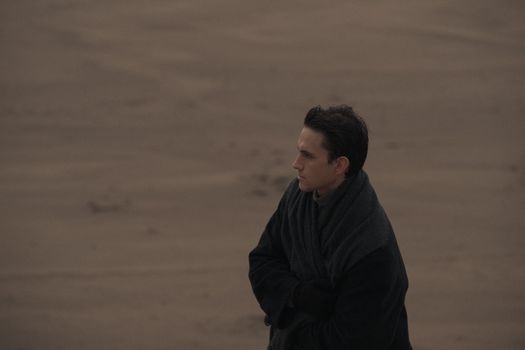 A man standing alone on a deserted beach, wrapped in a dark coat.