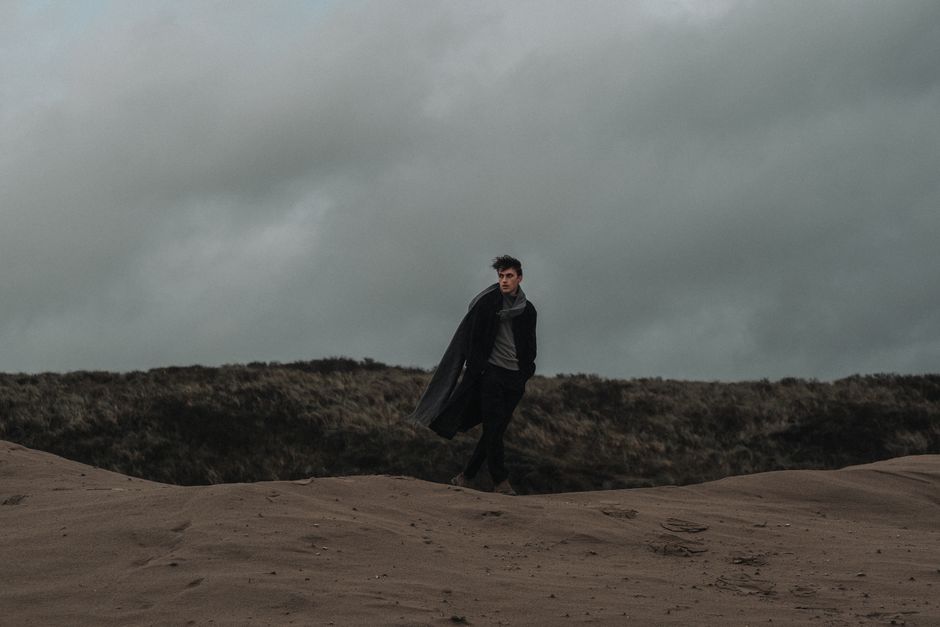 Stylish man in dark coat strides across a desolate sandy landscape under an overcast sky, exuding elegance.