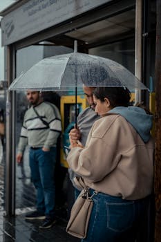 People gather under umbrellas at a rainy urban bus stop, capturing a moment of everyday life.