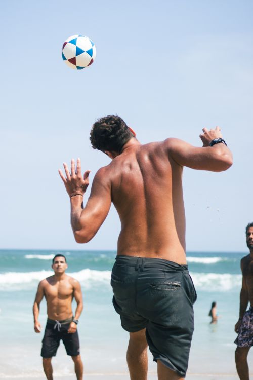 Free Three men enjoying a game of beach soccer by the ocean under a clear sky. Stock Photo