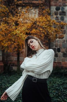 A fashionable woman in knitwear posing against a backdrop of autumn leaves and stone wall.
