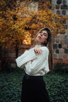 A stylish woman in a white sweater poses gracefully in a park with vibrant autumn foliage.