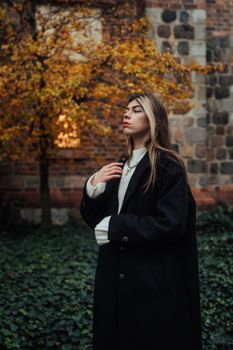 Stylish woman posing in autumn park with ivy and stone backdrop, exuding elegance.