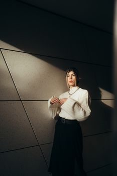 Fashionable woman poses stylishly in sunlight, wearing a white sweater and black skirt.