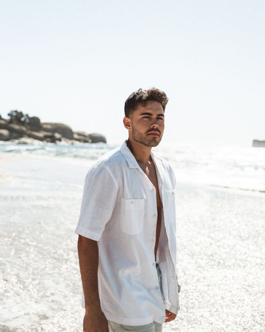 A confident man stands on a sunny beach wearing a white shirt, enjoying the coastal breeze.