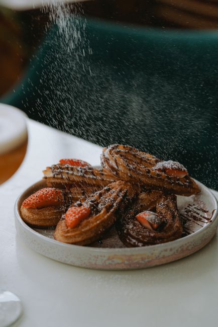 A bowl of golden, spiral-shaped fried pasta twists dusted heavily with cinnamon sugar, with a few scattered on a napkin beside it.