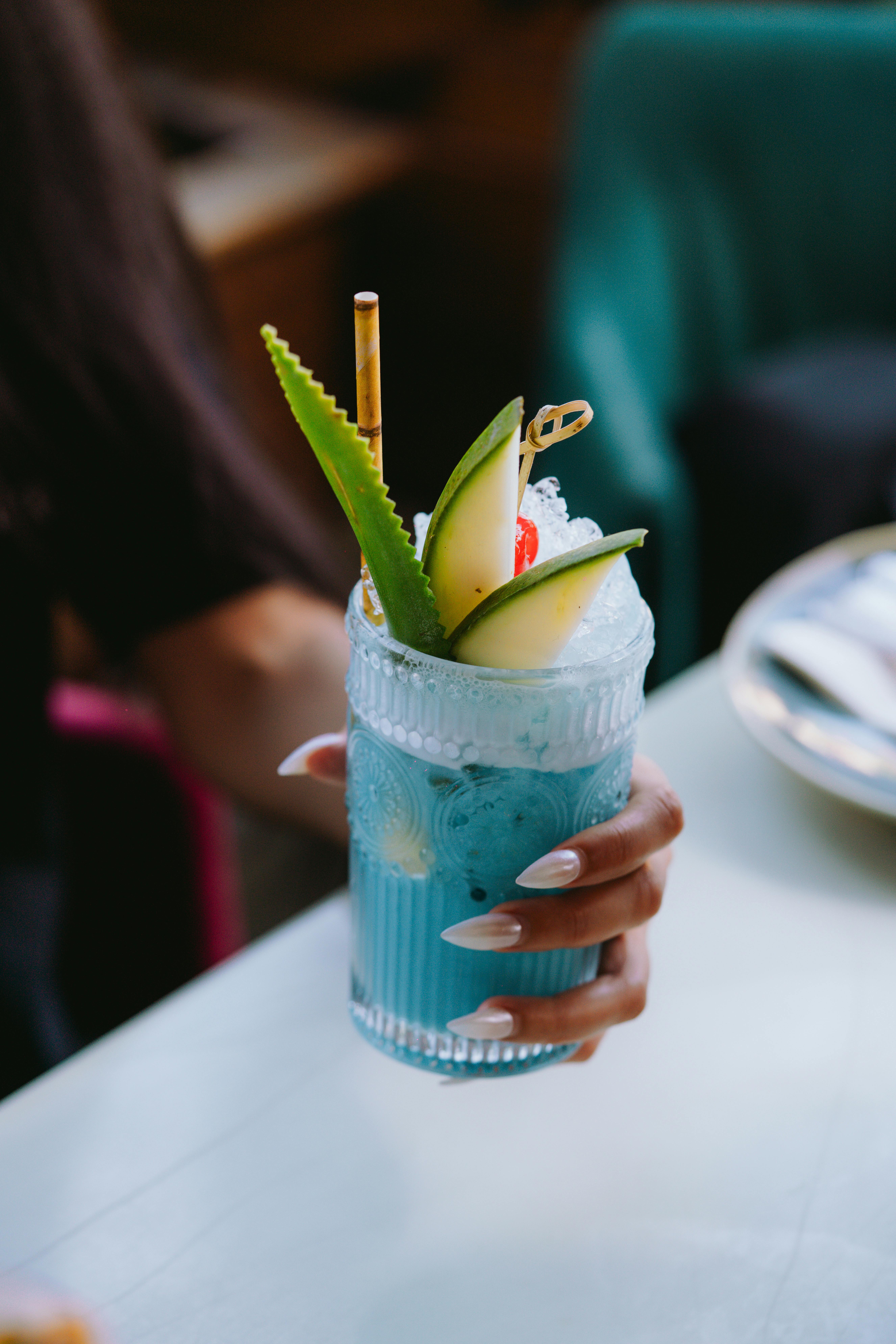 Free Close-up of a woman holding a vibrant blue cocktail garnished with fruit and ice in a glass. Stock Photo