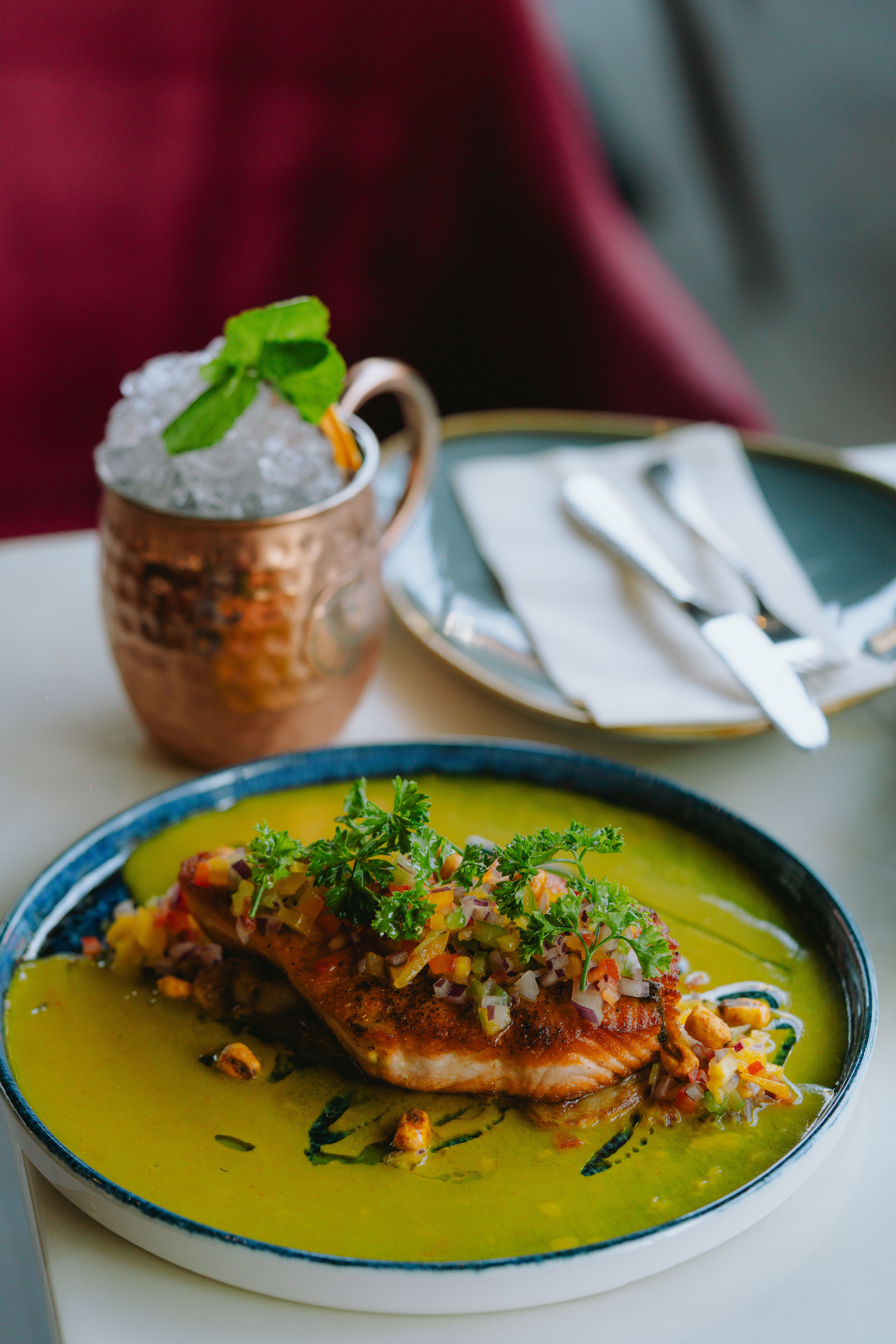 Elegant salmon dish with a vibrant green sauce, garnished with herbs and served with a refreshing cocktail.