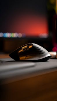 Close-up of an ergonomic wireless mouse on a desk with a blurred computer screen in the background.
