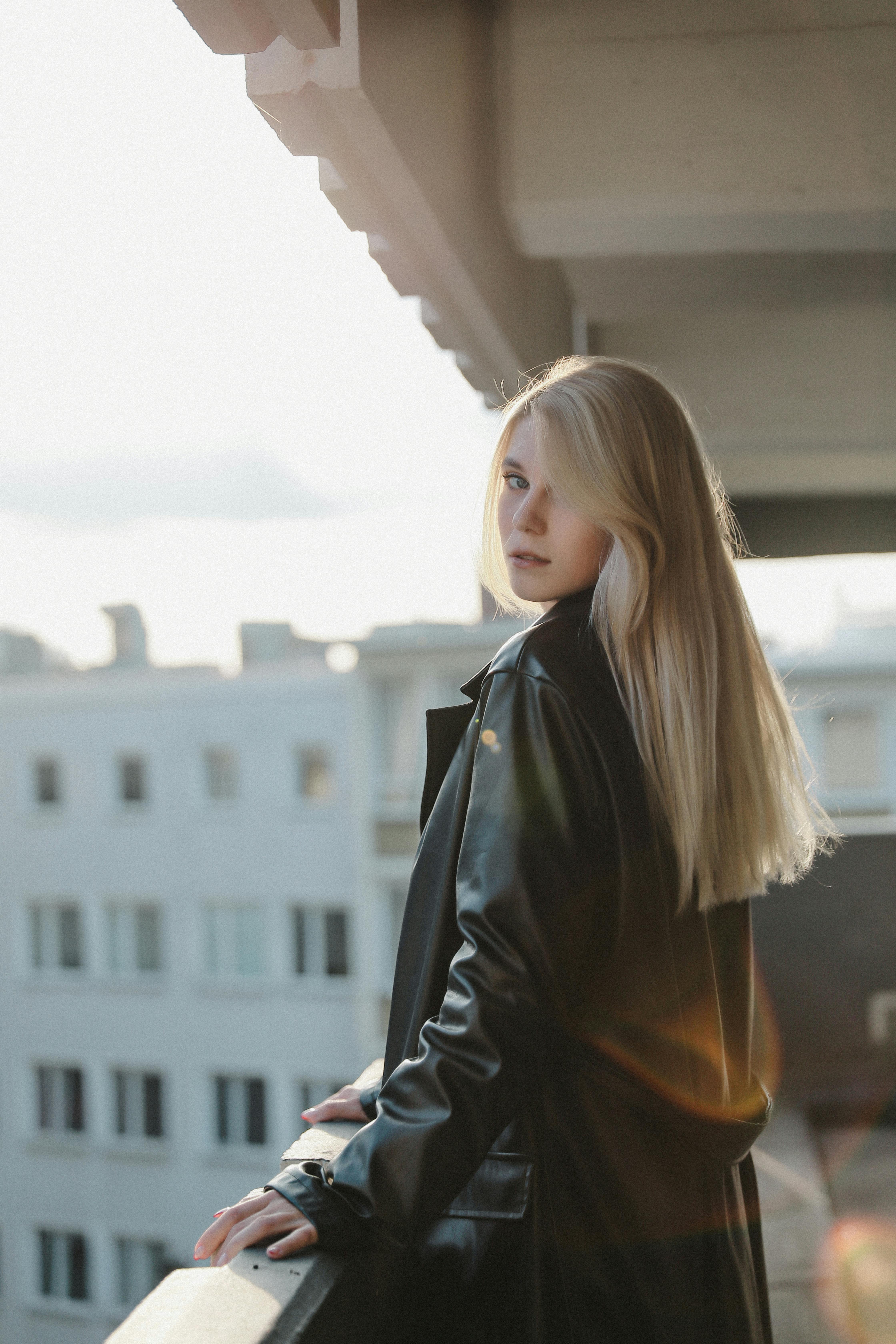 A fashionable woman in a leather coat standing on a balcony with city buildings in the background.