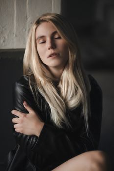 Elegant blonde woman with closed eyes posing in a black leather jacket indoors.