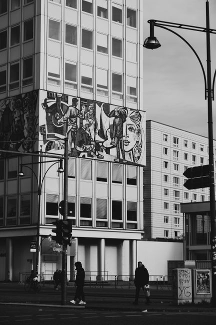 Black and white photo of a Berlin street with a striking mural and pedestrians.