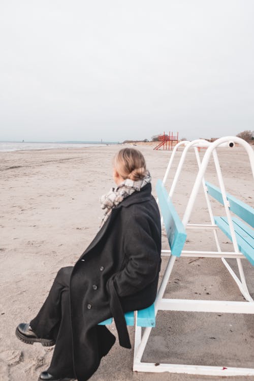Blonde Woman in Black Coat Sitting on Bench on Beach