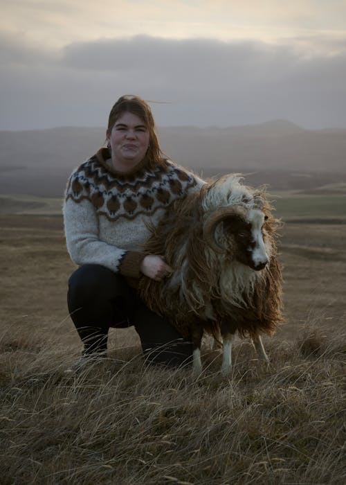 Woman in Warm Sweater Posing with a Sheep on a Pasture