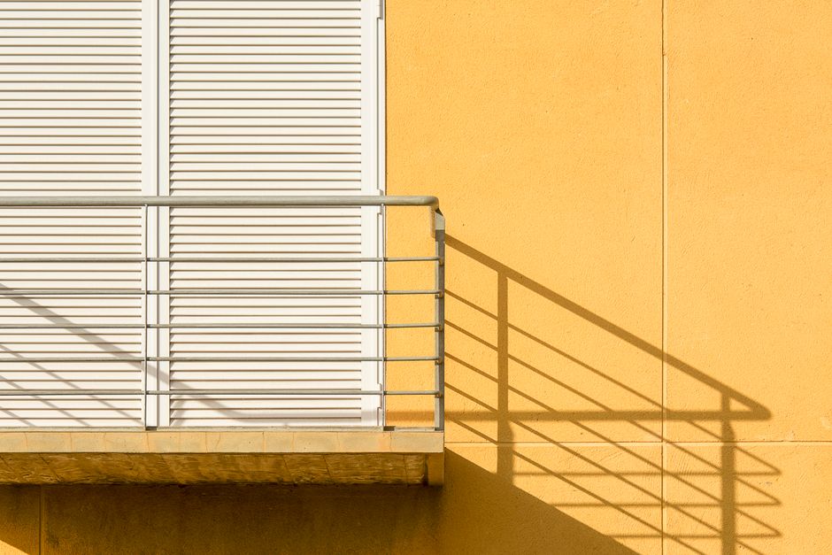 Minimalistic balcony railing casting a shadow on a vibrant yellow wall with shutters.