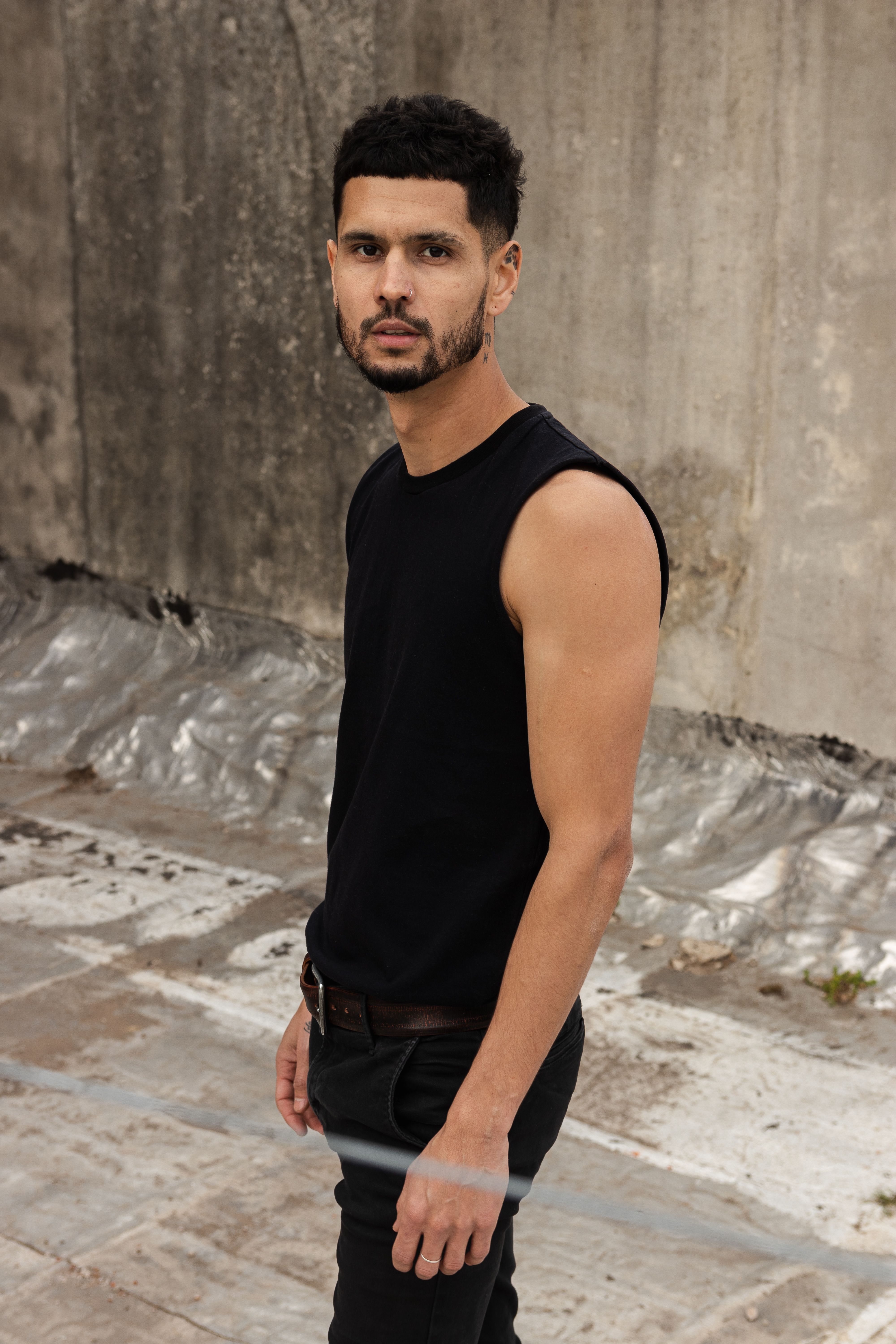 Portrait of a bearded man in a sleeveless top, posing against a city wall.