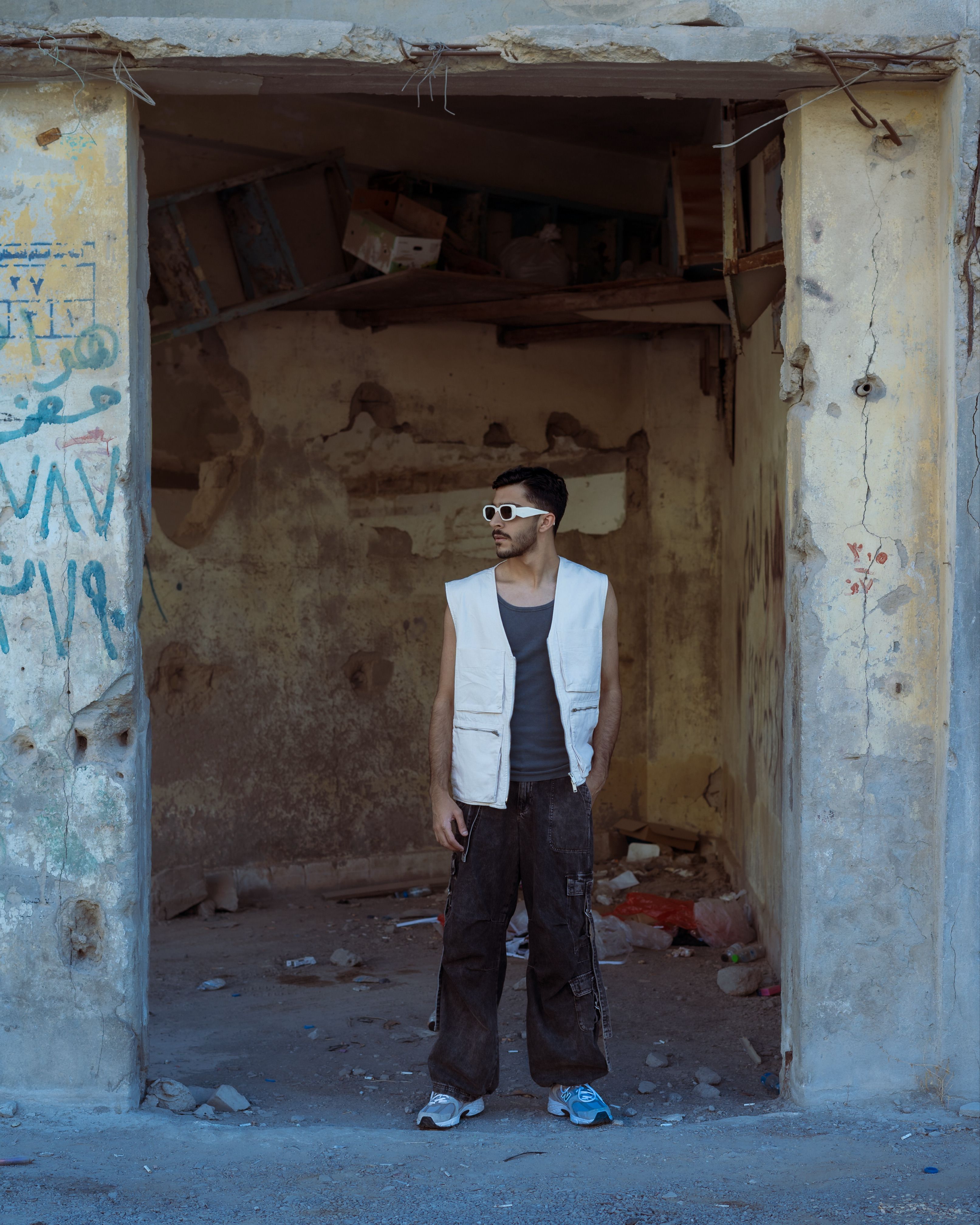 A stylish young man in sunglasses stands confidently in an abandoned urban setting, showcasing modern street style.