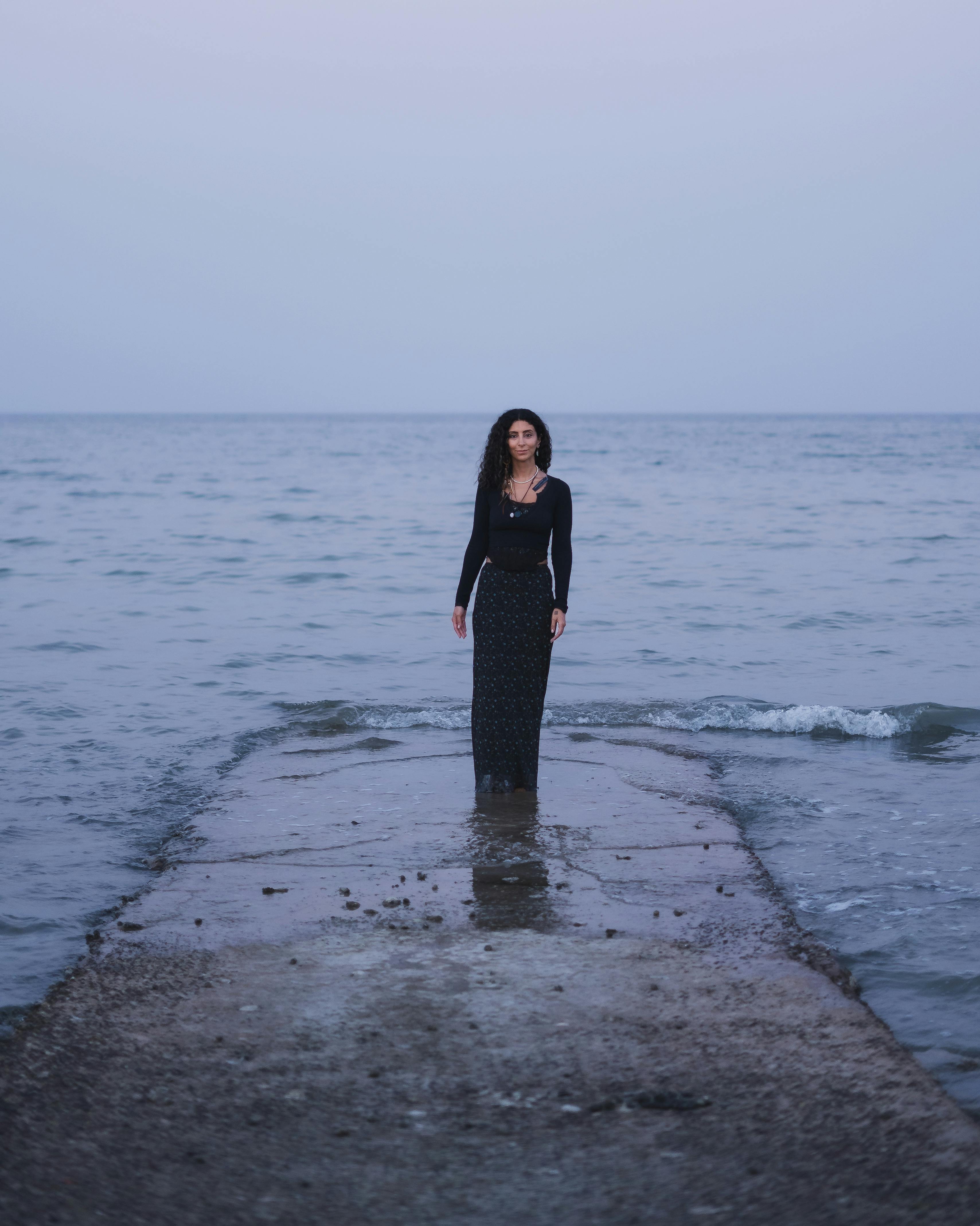 A woman in a black gown stands on a pier over shallow water under an overcast sky.