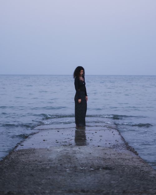 Free A woman stands alone on a pier, facing the calm sea during twilight, creating a serene atmosphere. Stock Photo