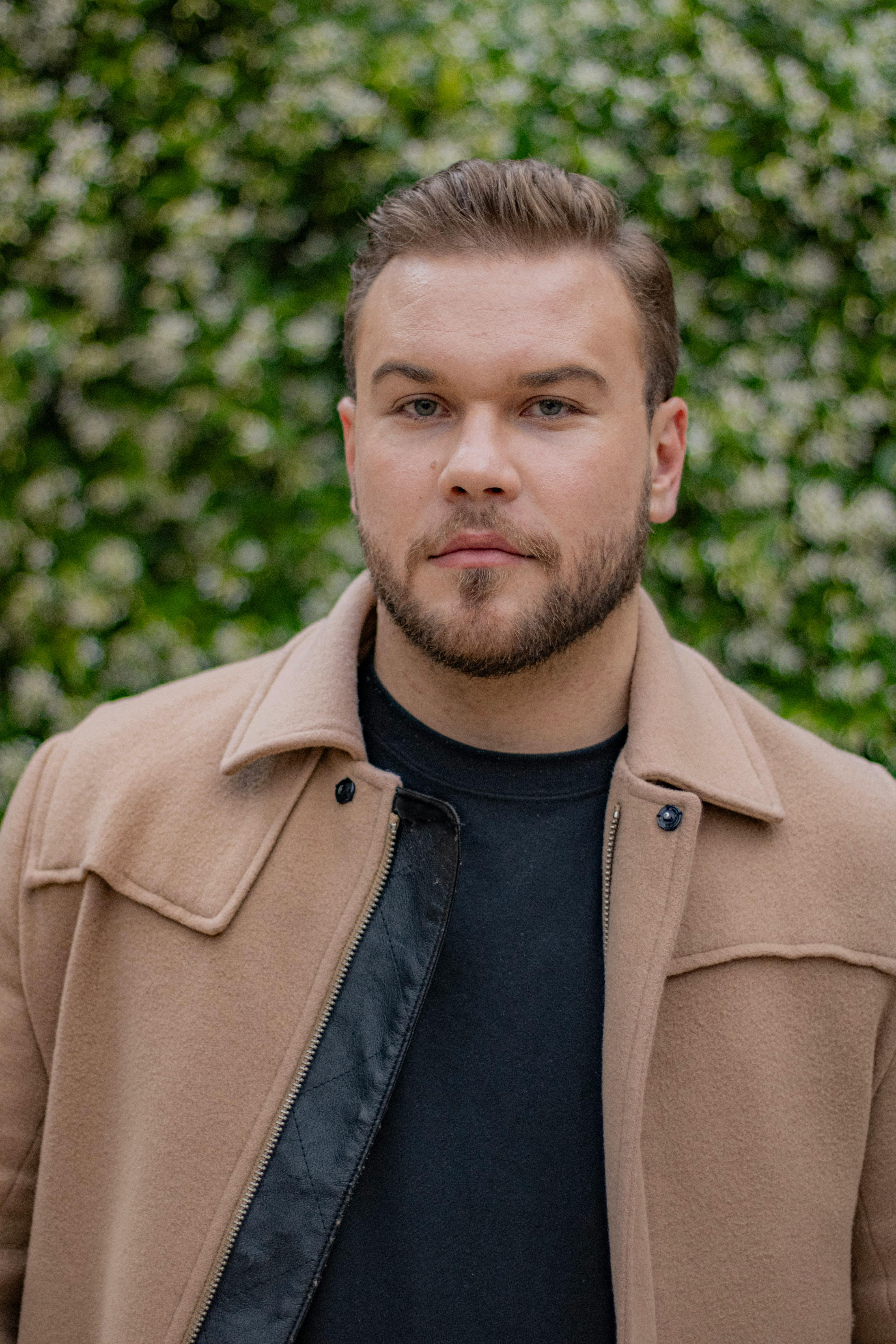 Portrait of a bearded man wearing a beige jacket against a leafy background.