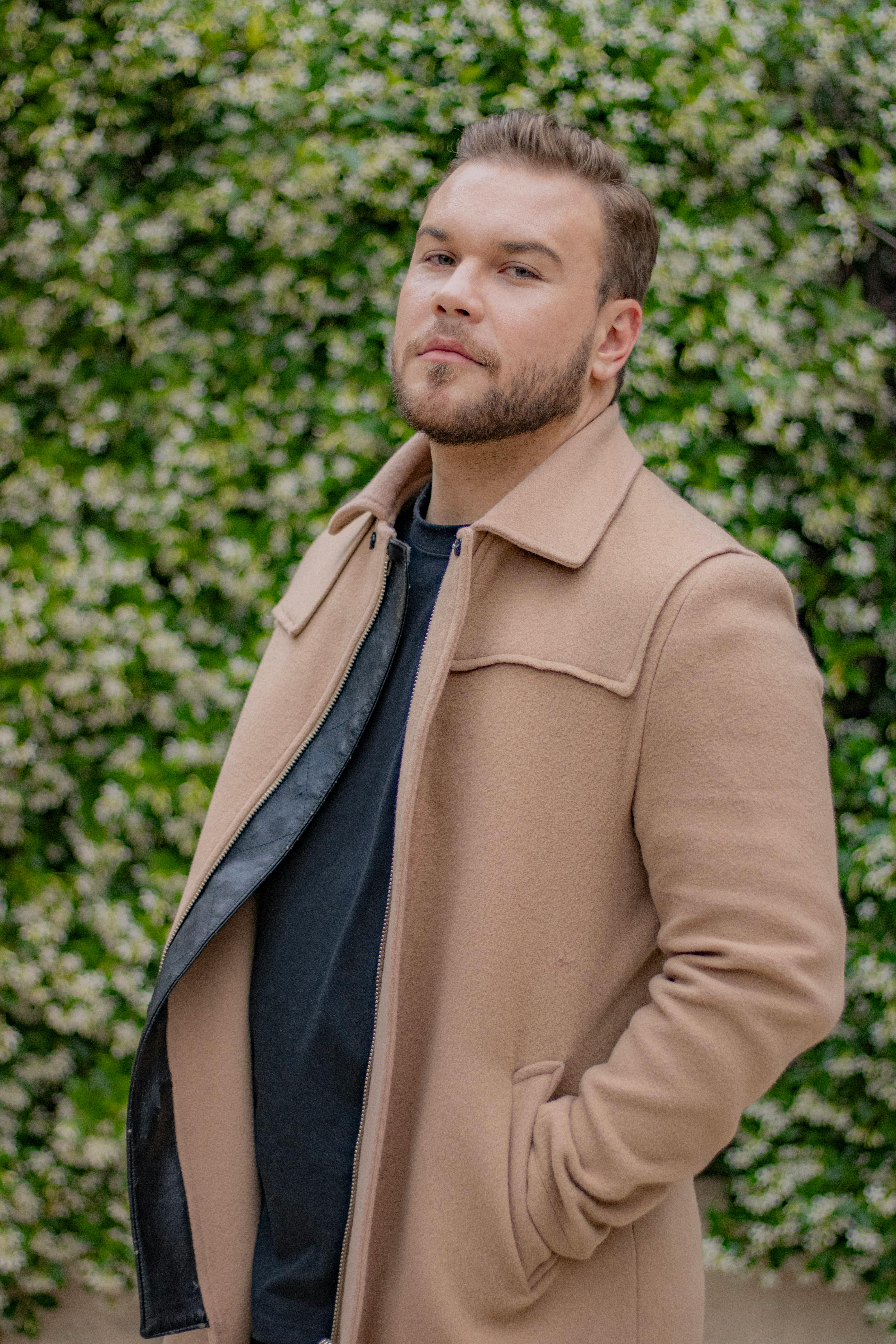 Man in a beige coat posing outdoors against a green leafy backdrop.