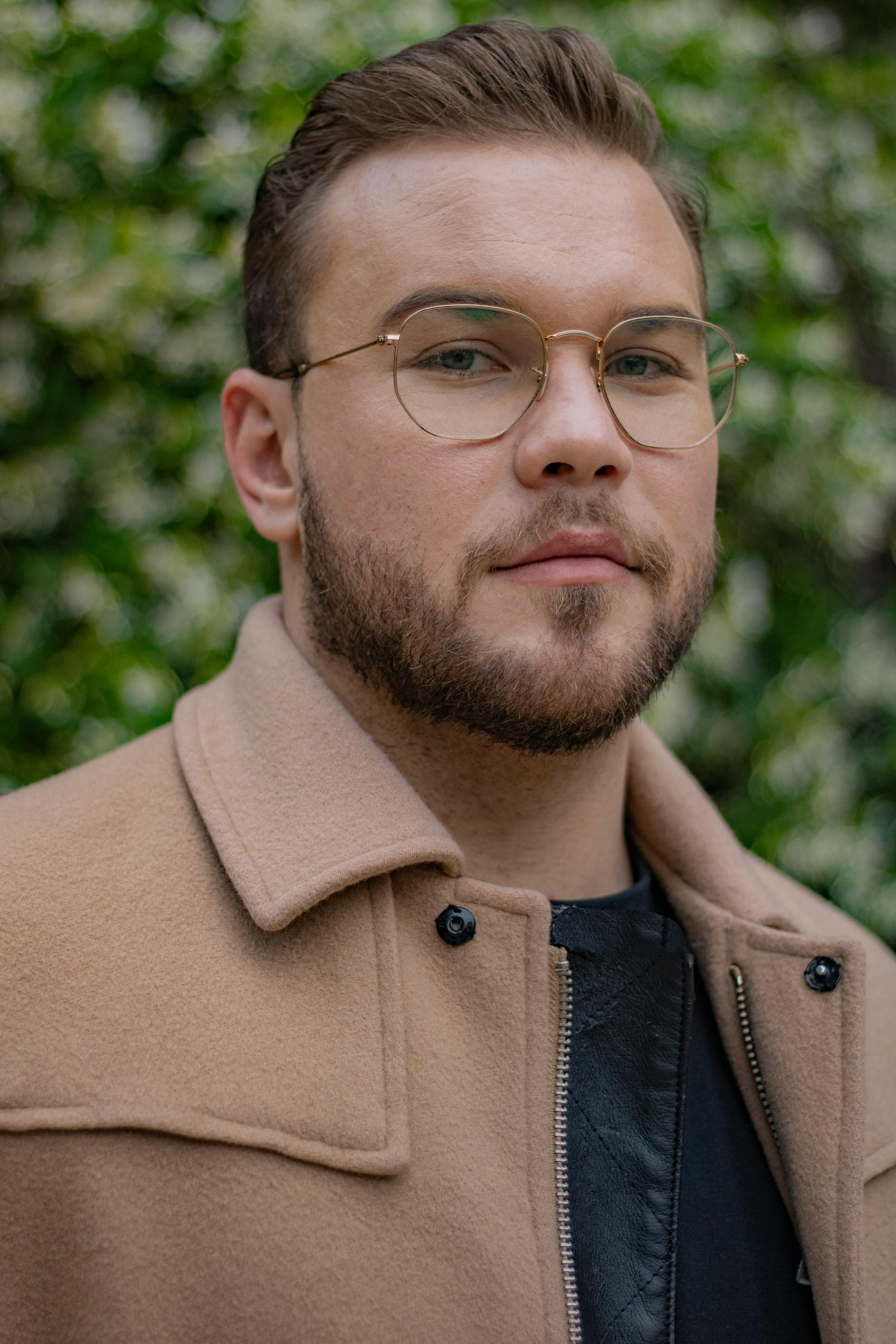 Fashionable young man with glasses and beard posing outdoors in a beige coat.