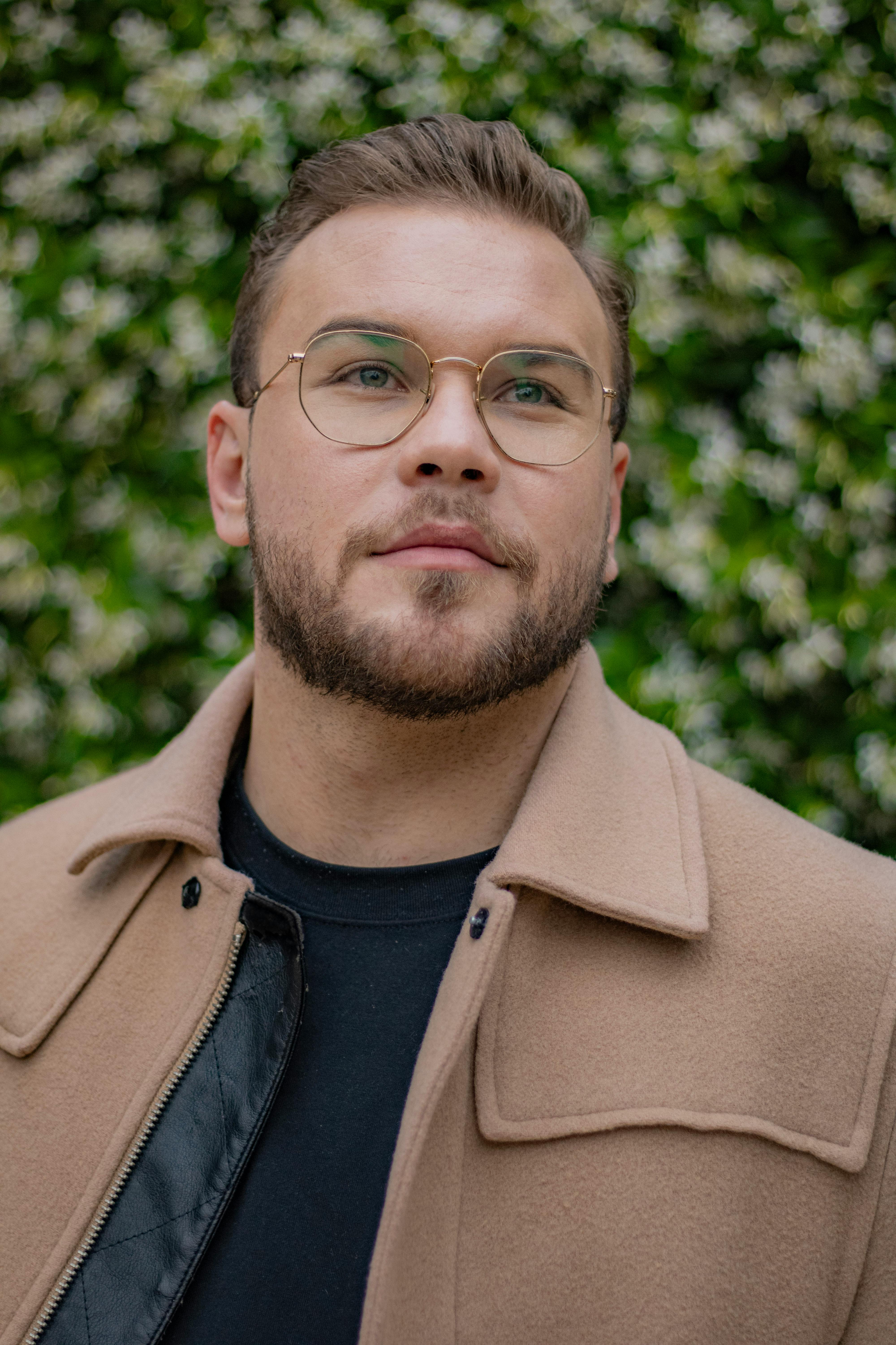 Portrait of a young man with eyeglasses and beard posing outdoors in a beige coat.