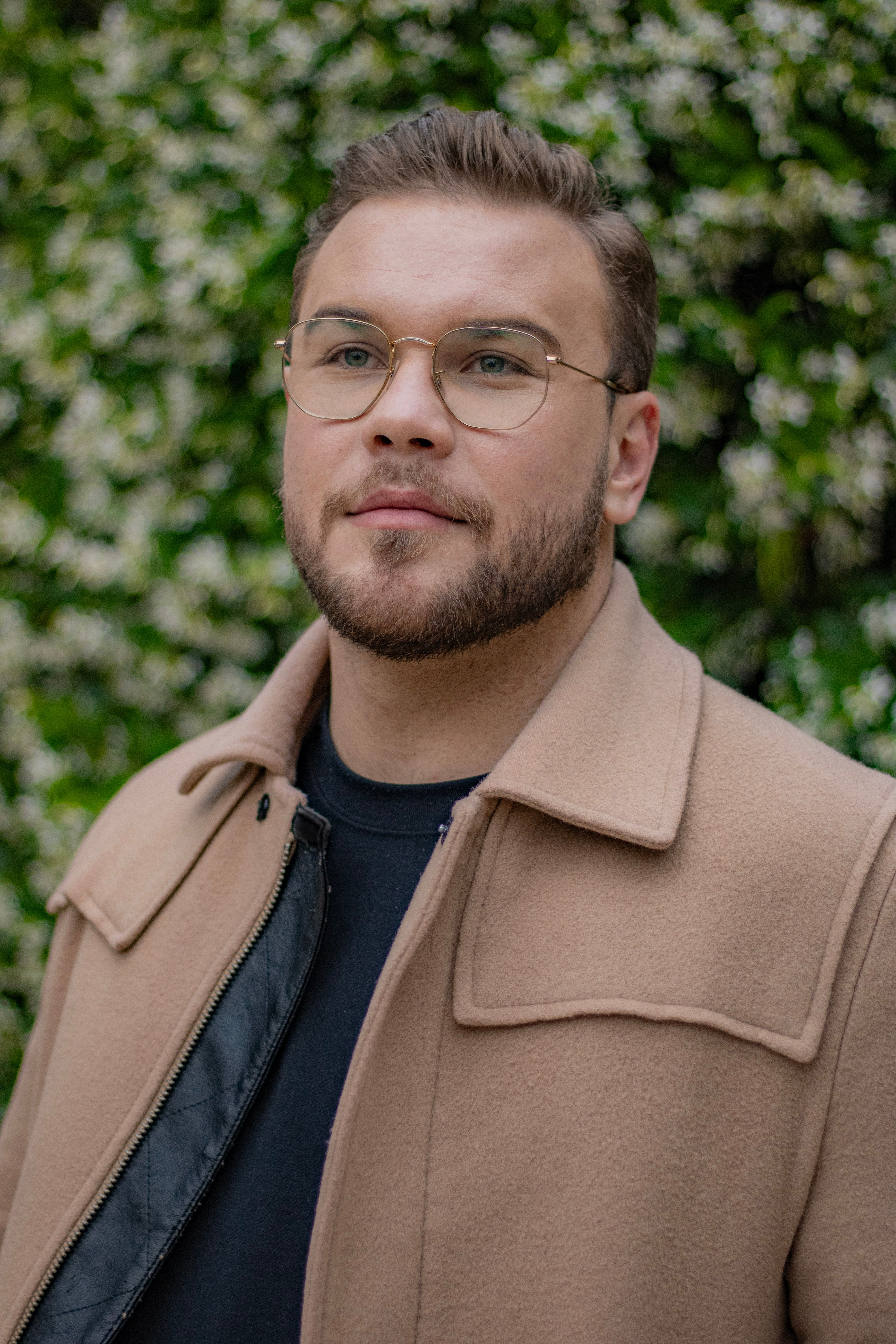 A man stands confidently in a beige coat and glasses against a lush green background.