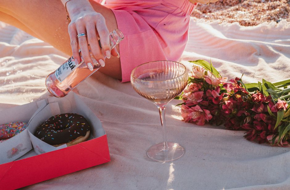A woman enjoys a summer beach picnic with donuts, flowers, and a refreshing drink.