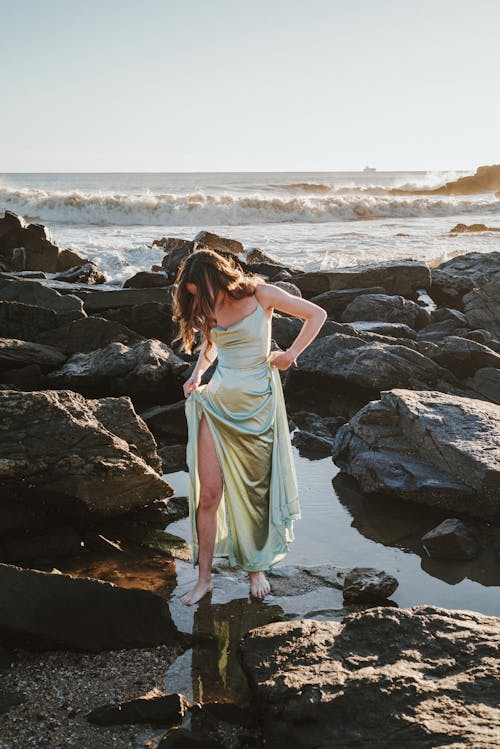 Woman in Long Gown Walking through Shallow Water between Rocks on Sea Shore