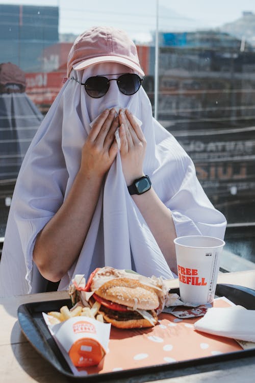 Person in a Ghost Costume and Sunglasses Sitting at the Table and Eating Fast Food 