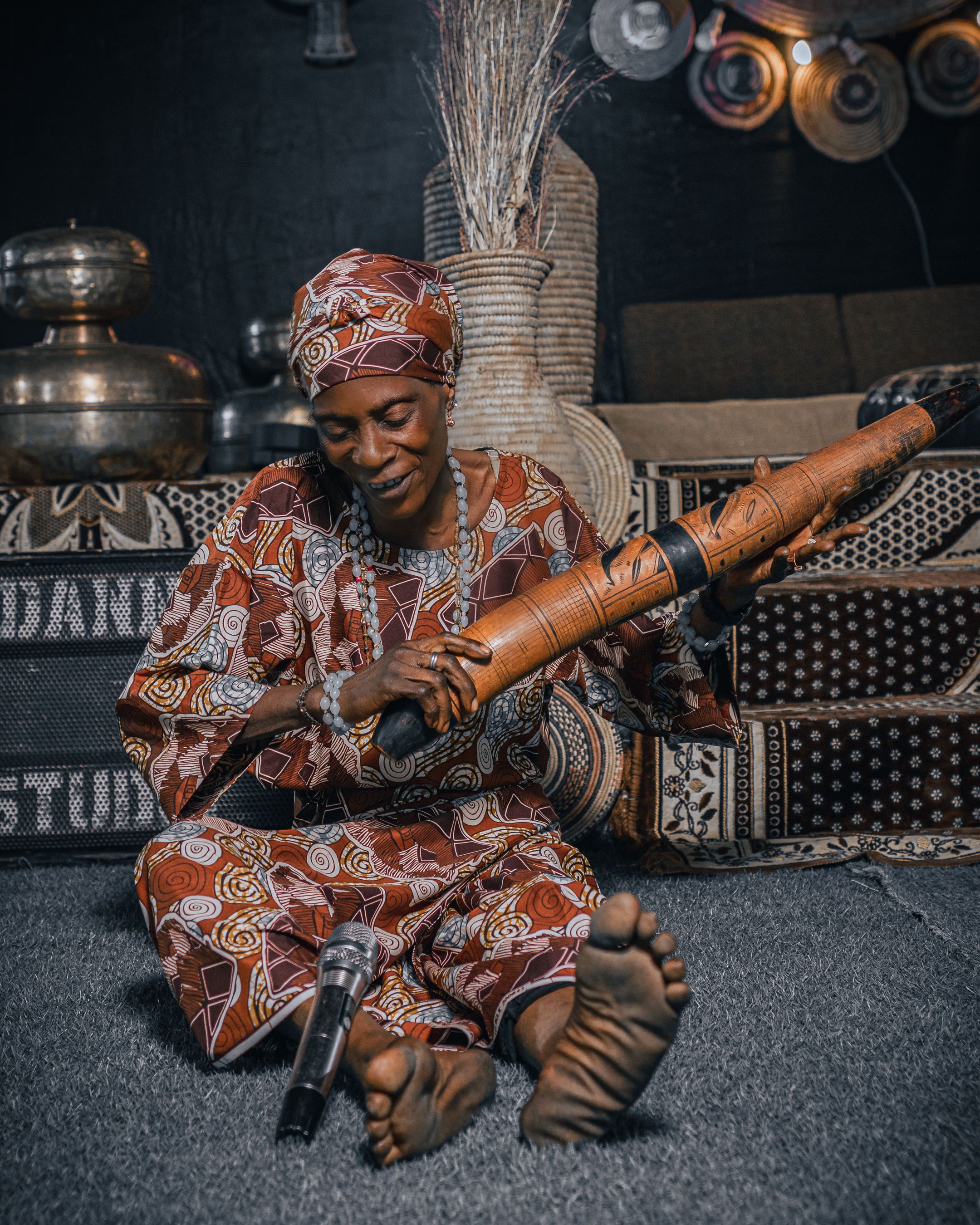 Smiling woman in patterned attire playing a traditional instrument indoors, sitting on a carpet.