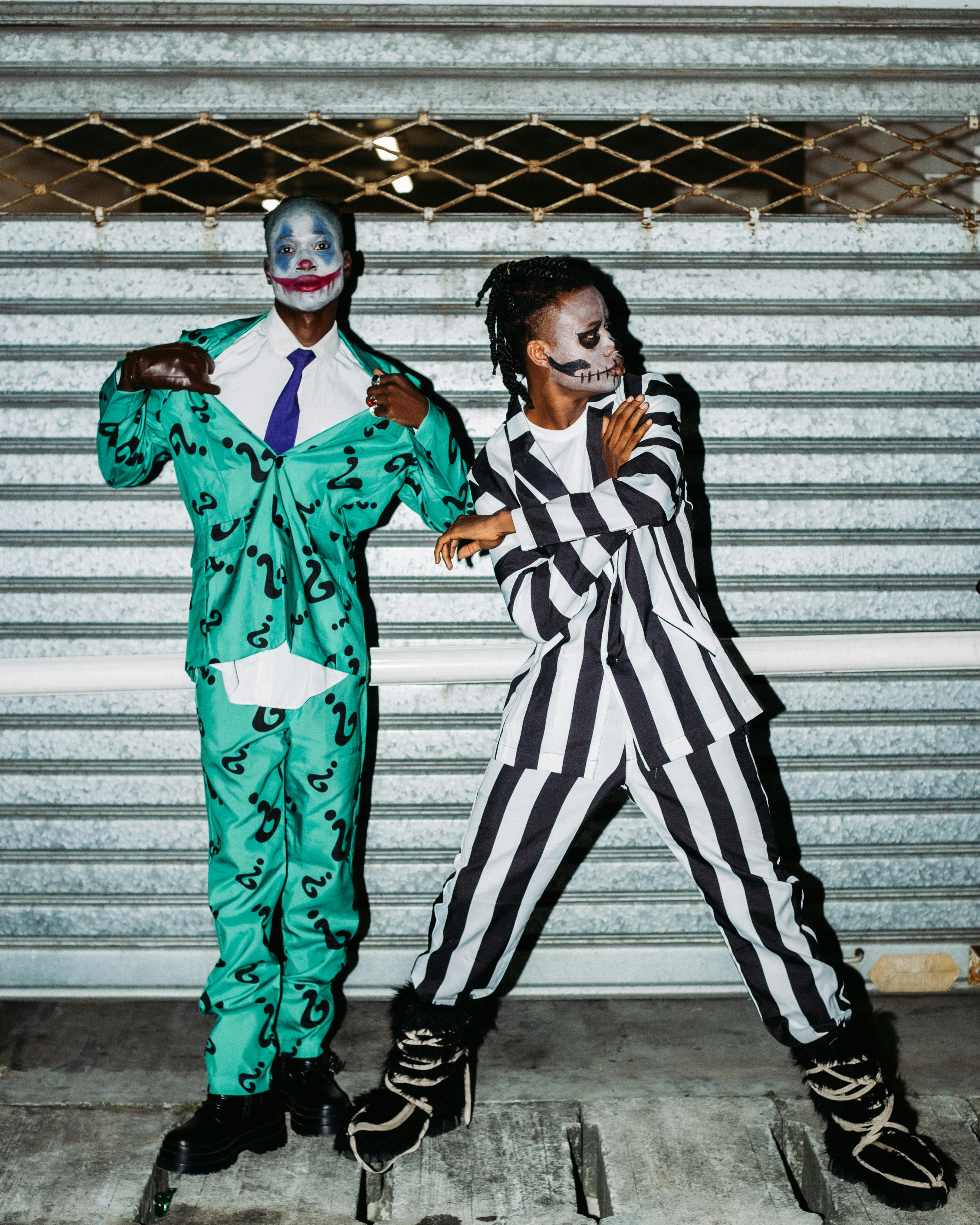 Two people in bold Halloween costumes and makeup posing creatively against a metal backdrop.