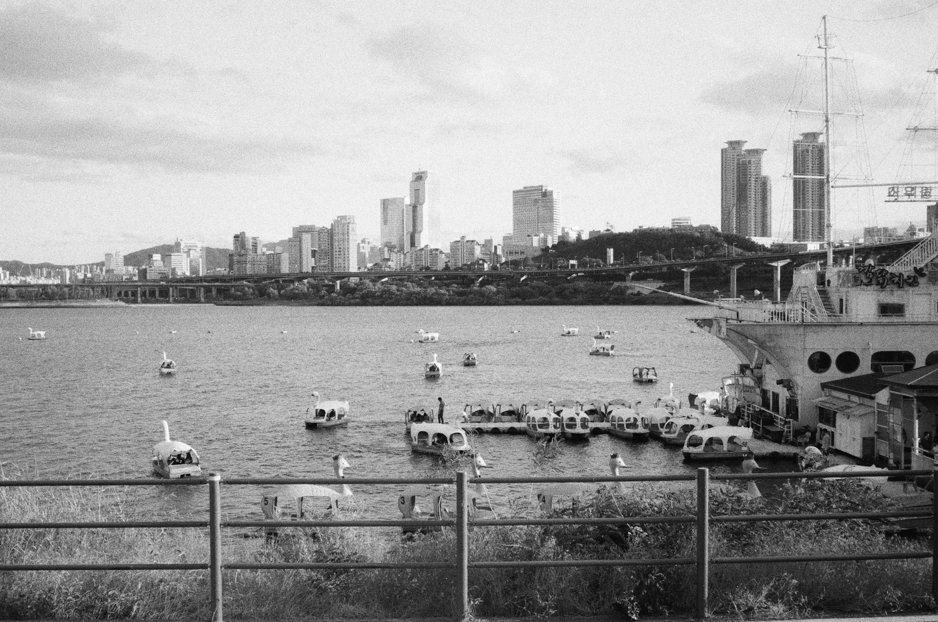 Black and white cityscape of Seoul with boats on a river and skyscrapers in the background.