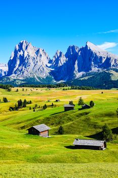 Serene view of the Dolomites mountains and lush meadows, featuring rustic huts under a clear blue sky.