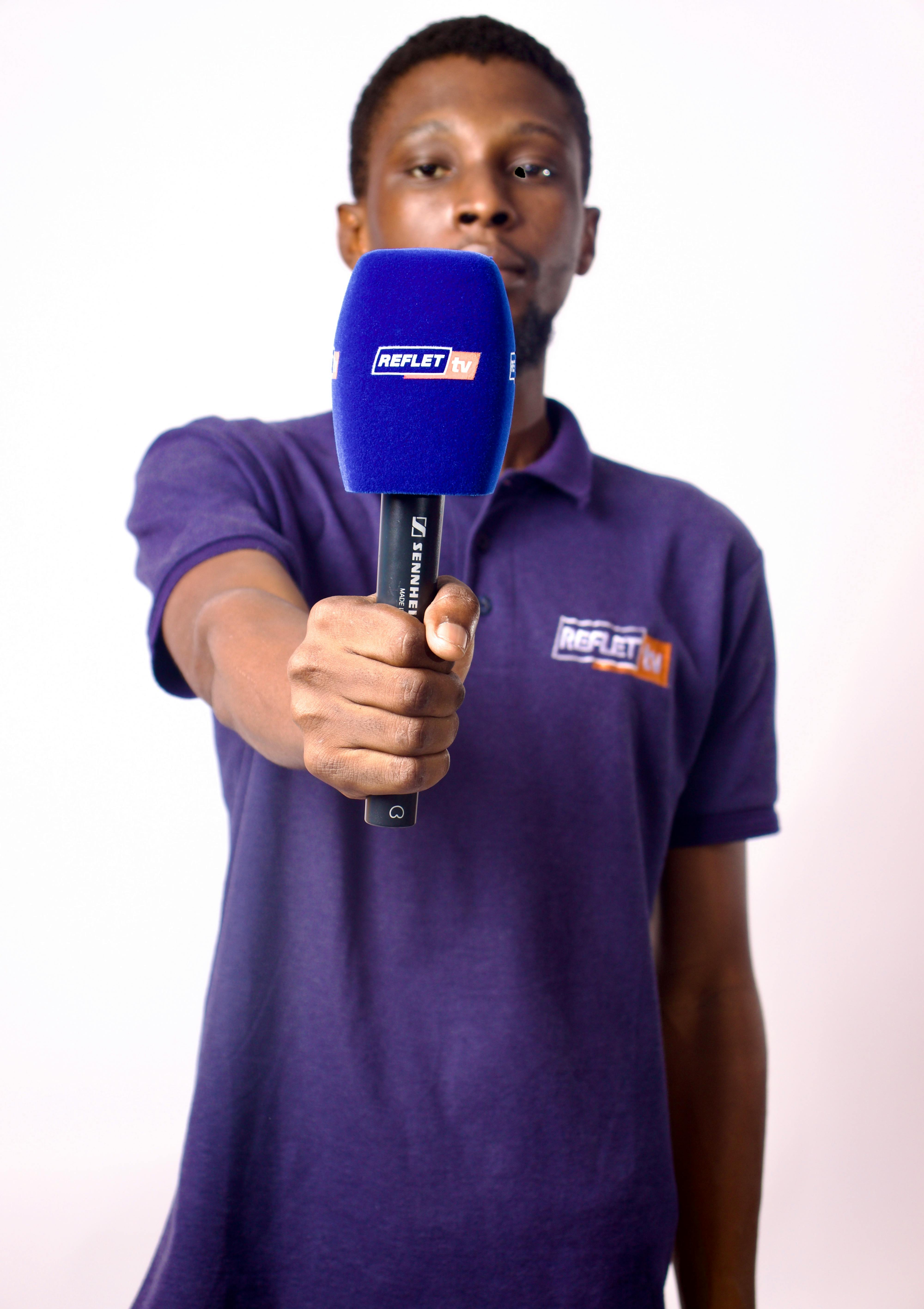 A young African reporter in a studio holding a microphone, wearing a purple shirt.