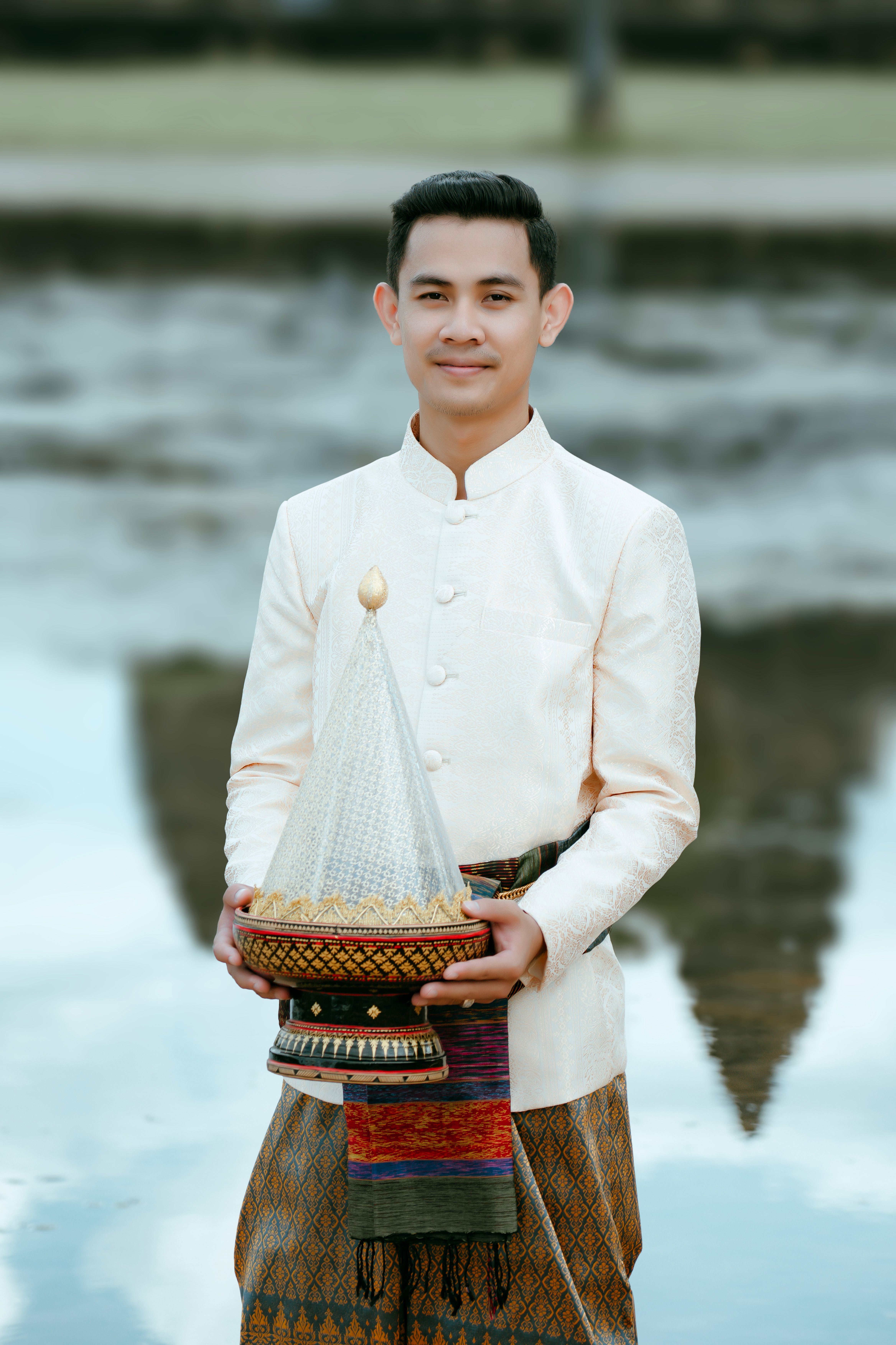 Man in traditional attire holding a ceremonial object with Angkor Wat reflection in the background.