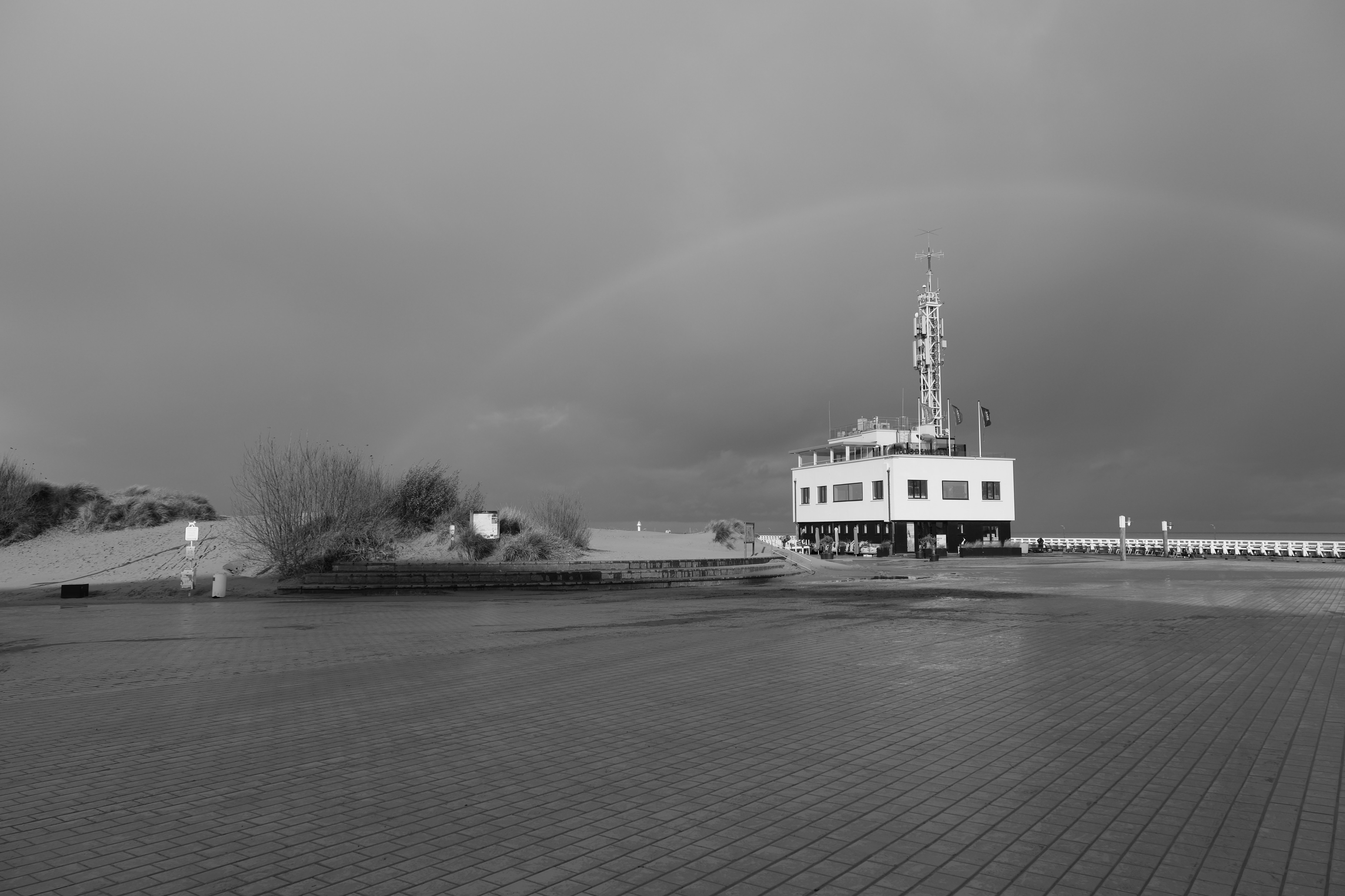 Black and white coastal landscape featuring a modern building and empty square under dramatic sky.
