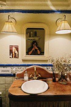 Warm-lit bathroom with a woman taking a mirror selfie, featuring decor and lights.