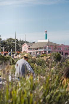 A vibrant coastal scene in Monterey, CA, featuring a lighthouse, pink building, and people enjoying a sunny day.