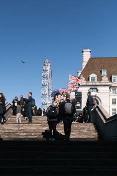 Tourists walking up stairs near the iconic London Eye on a clear day with Union Jack flags visible.