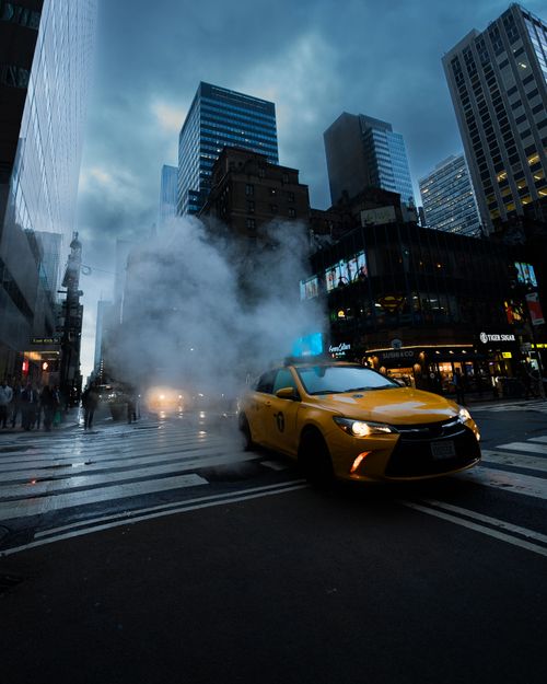 Free A yellow taxi drives through a bustling New York City street surrounded by skyscrapers and evening lights. Stock Photo