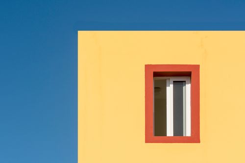 Free A simple and vibrant minimalist architecture shot of a yellow building facade with a red-trimmed window against a clear blue sky. Stock Photo